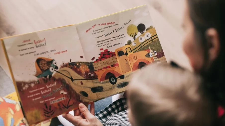 Bilden är ett fotografi av en vuxen person med ett barn i knäet som läser en bok. 