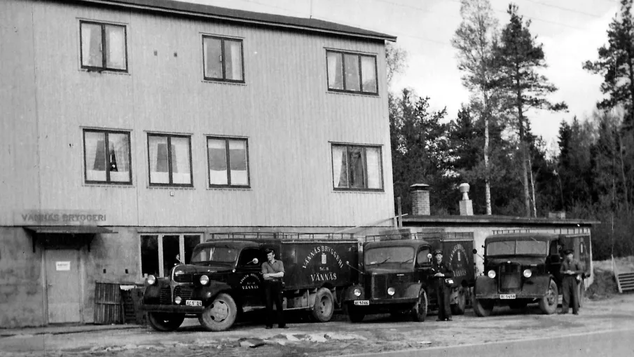 Ett gammalt svartvitt fotografi av Vännäs Bryggeri, men tre av företagets fordon och förare parkerade framför byggnaden.