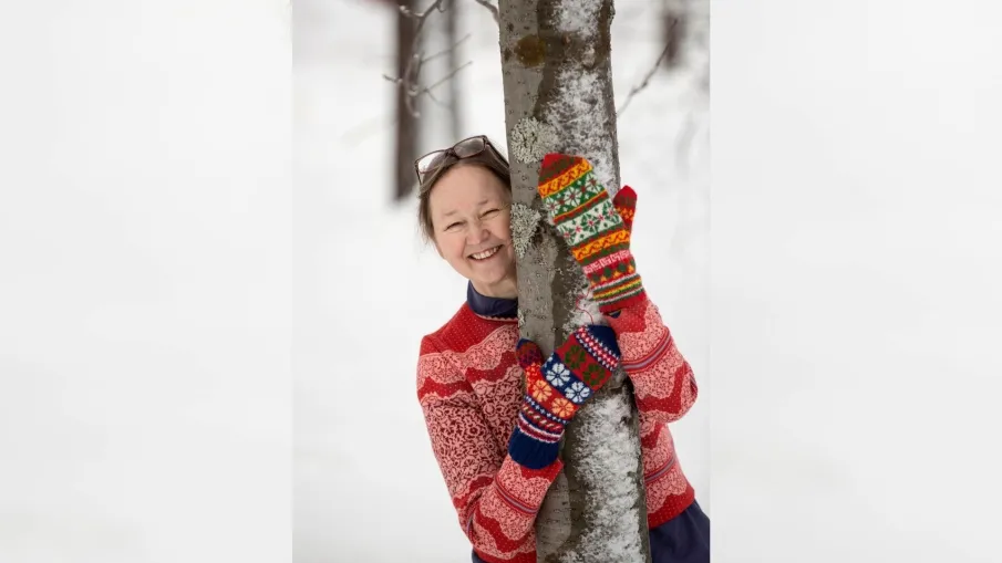 Fotografi av Erika Nordvall Falck med armarna omkring ett träd och färgglada stickade vantar på händerna.